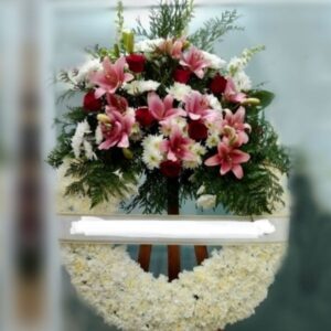Corona de flores funerarias de margaritas blancas con penacho de azucenas en tonos rosas.